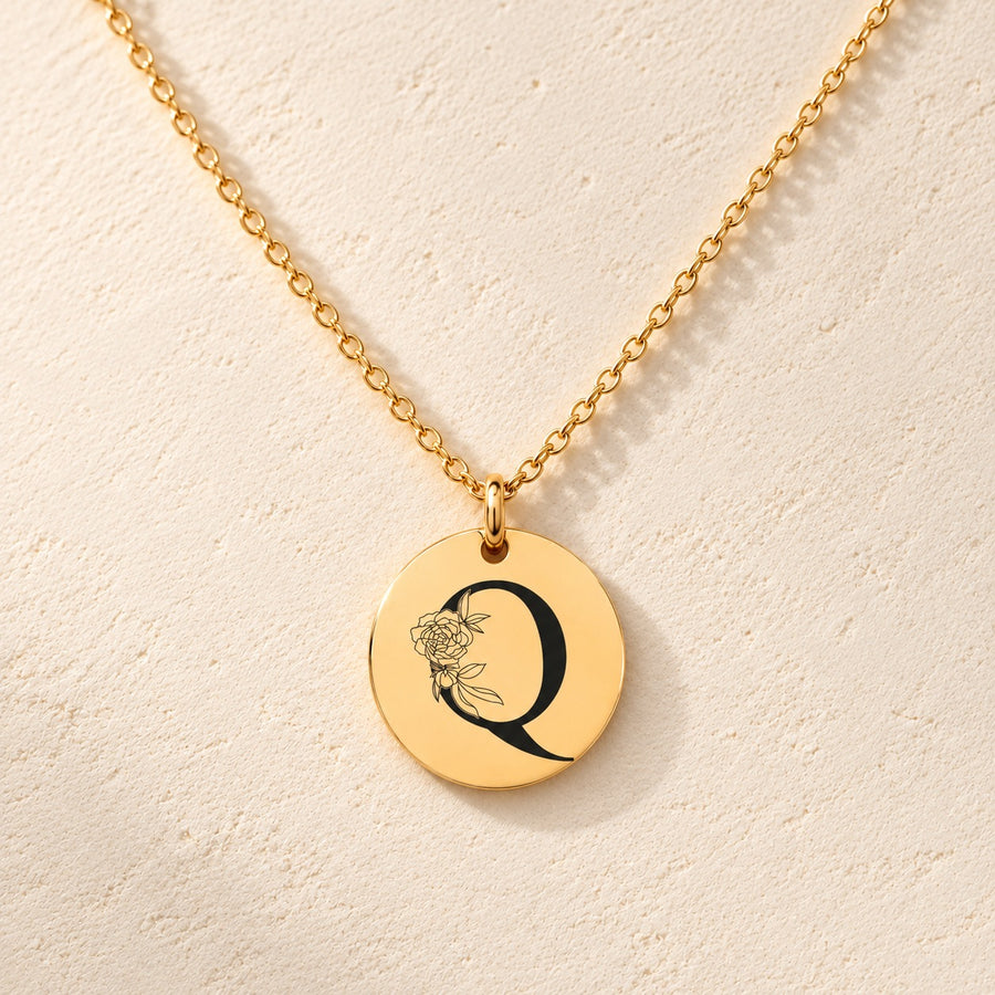 Collier initiale florale Q personnalisé doré, médaille ronde gravée avec initiale Q et fleurs, sur fond ivoire.