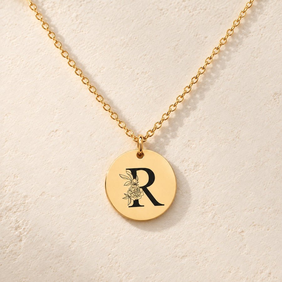 Collier initiale florale R personnalisé doré, médaille ronde gravée avec initiale R et fleurs, sur fond ivoire.