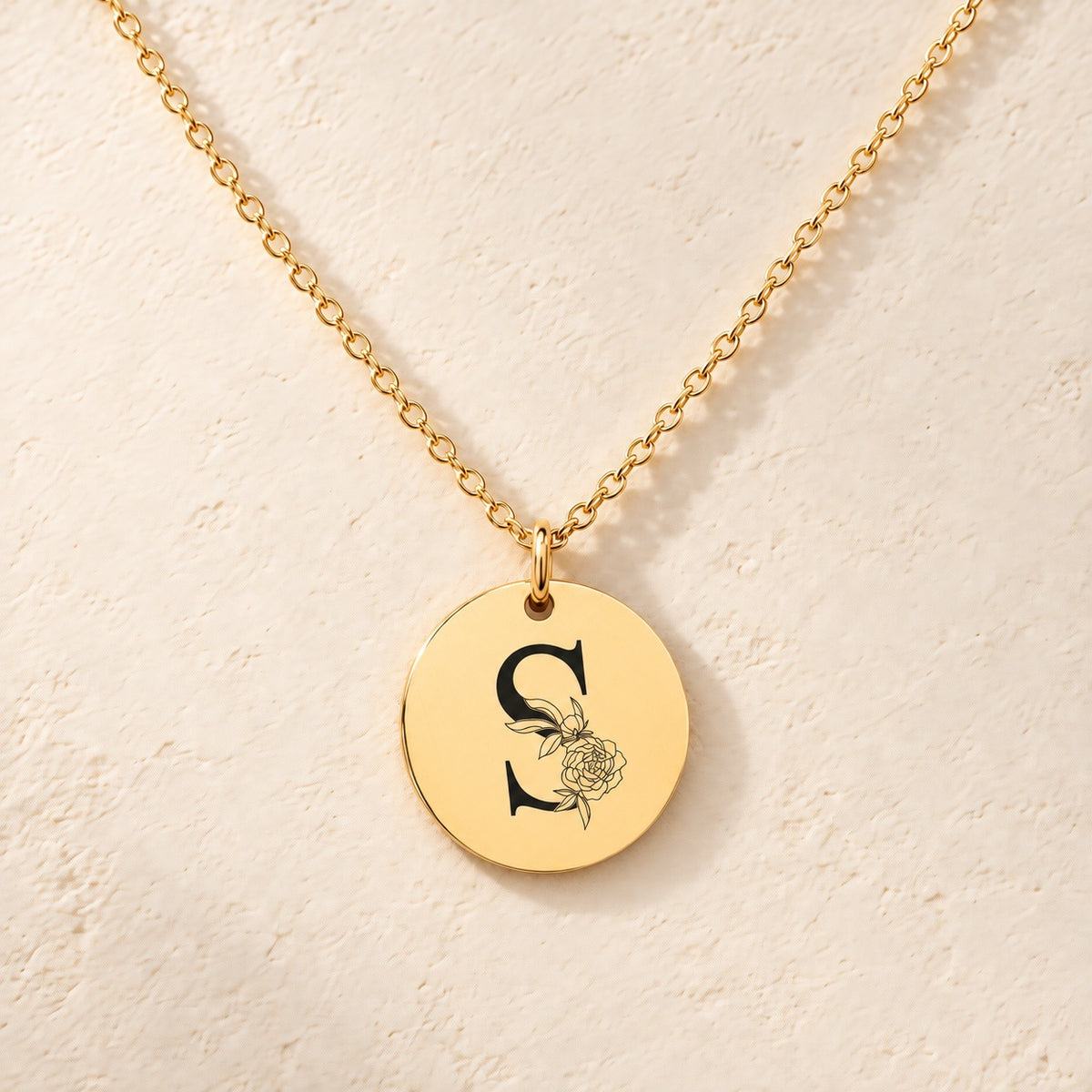 Collier initiale florale S personnalisé doré, médaille ronde gravée avec initiale S et fleurs, sur fond ivoire.