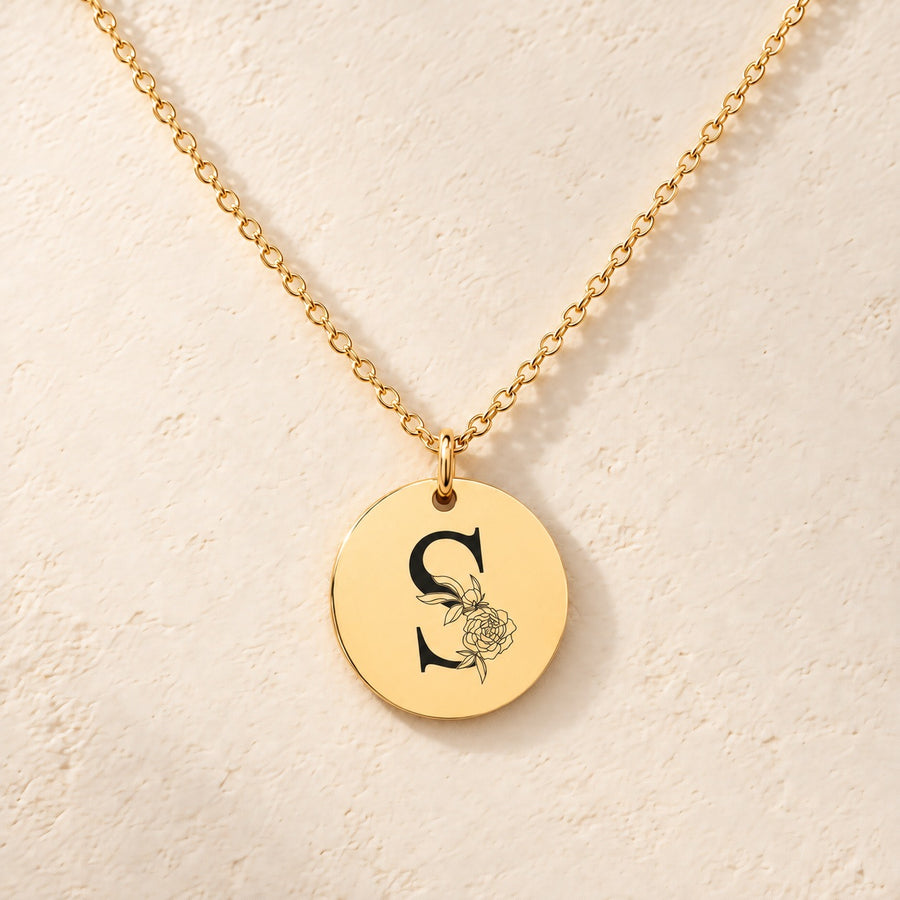 Collier initiale florale S personnalisé doré, médaille ronde gravée avec initiale S et fleurs, sur fond ivoire.