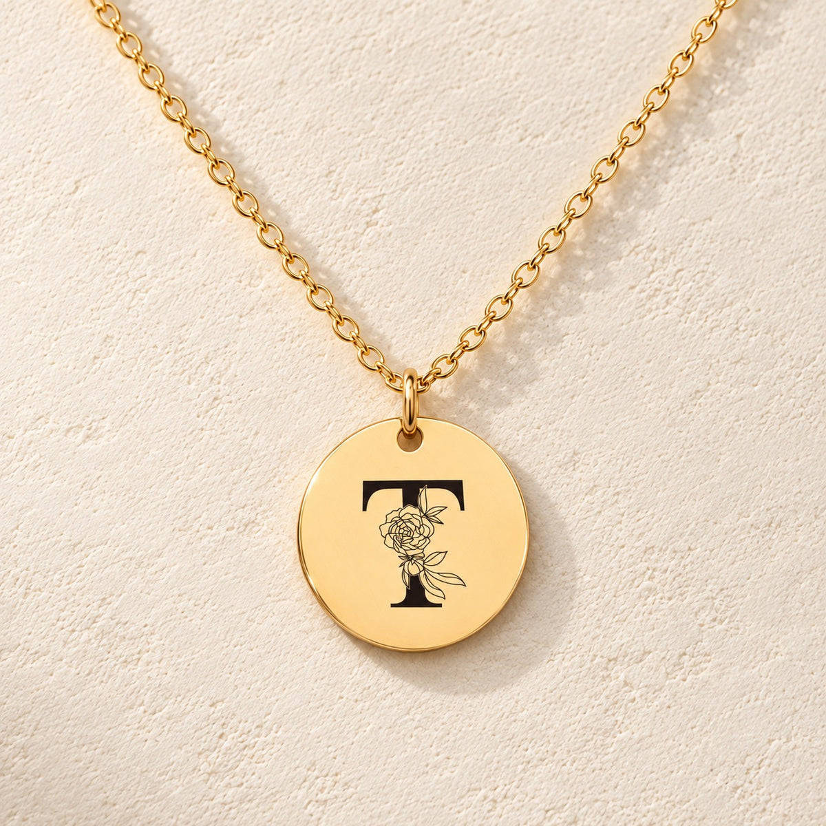 Collier initiale florale T personnalisé doré, médaille ronde gravée avec initiale T et fleurs, sur fond ivoire.