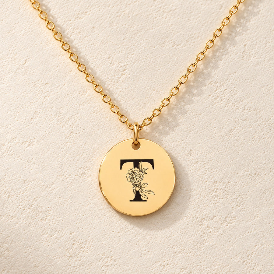 Collier initiale florale T personnalisé doré, médaille ronde gravée avec initiale T et fleurs, sur fond ivoire.