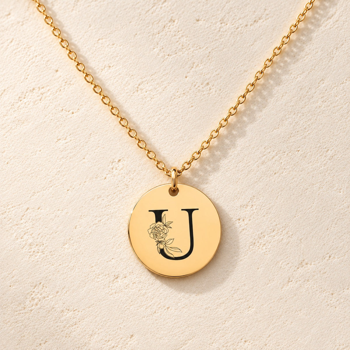 Collier initiale florale U personnalisé doré, médaille ronde gravée avec initiale U et fleurs, sur fond ivoire.