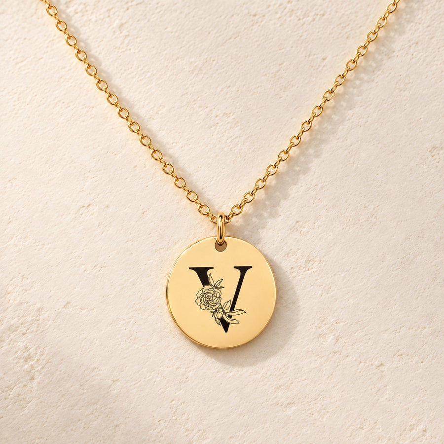 Collier initiale florale V personnalisé doré, médaille ronde gravée avec initiale V et fleurs, sur fond ivoire.