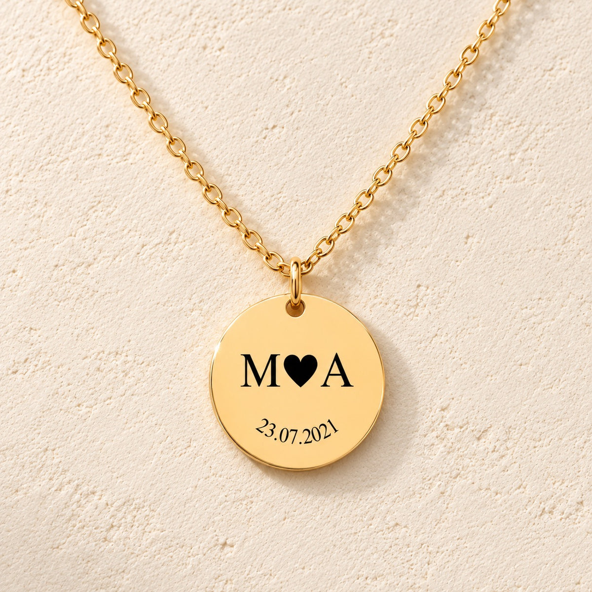 Collier initiales entrelacées Etched in Time personnalisé doré, médaille ronde gravée avec initiales M et A et date 23.07.2021, sur fond ivoire.