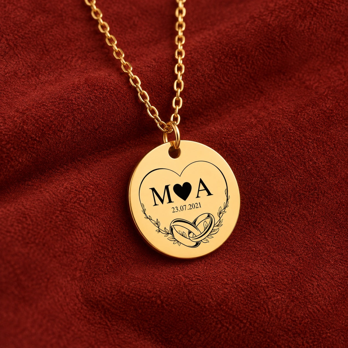 Médaille ronde dorée avec initiales M et A, deux alliances et date 23.07.2021, sur suédine rouge sanguine.