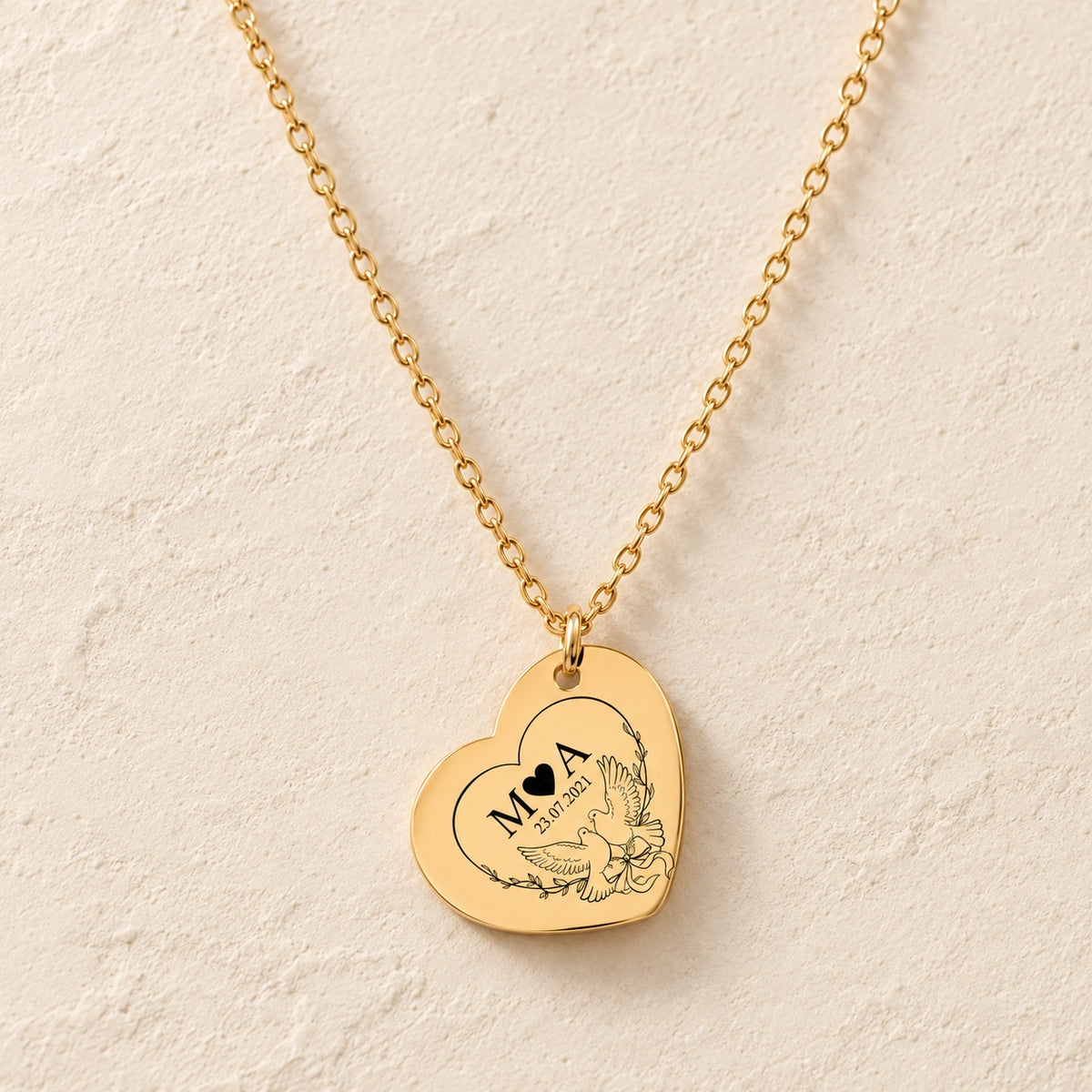 Collier Eternal Love doré, pendentif en forme de cœur avec deux colombes, initiales M et A et date 23.07.2021 sur fond ivoire.