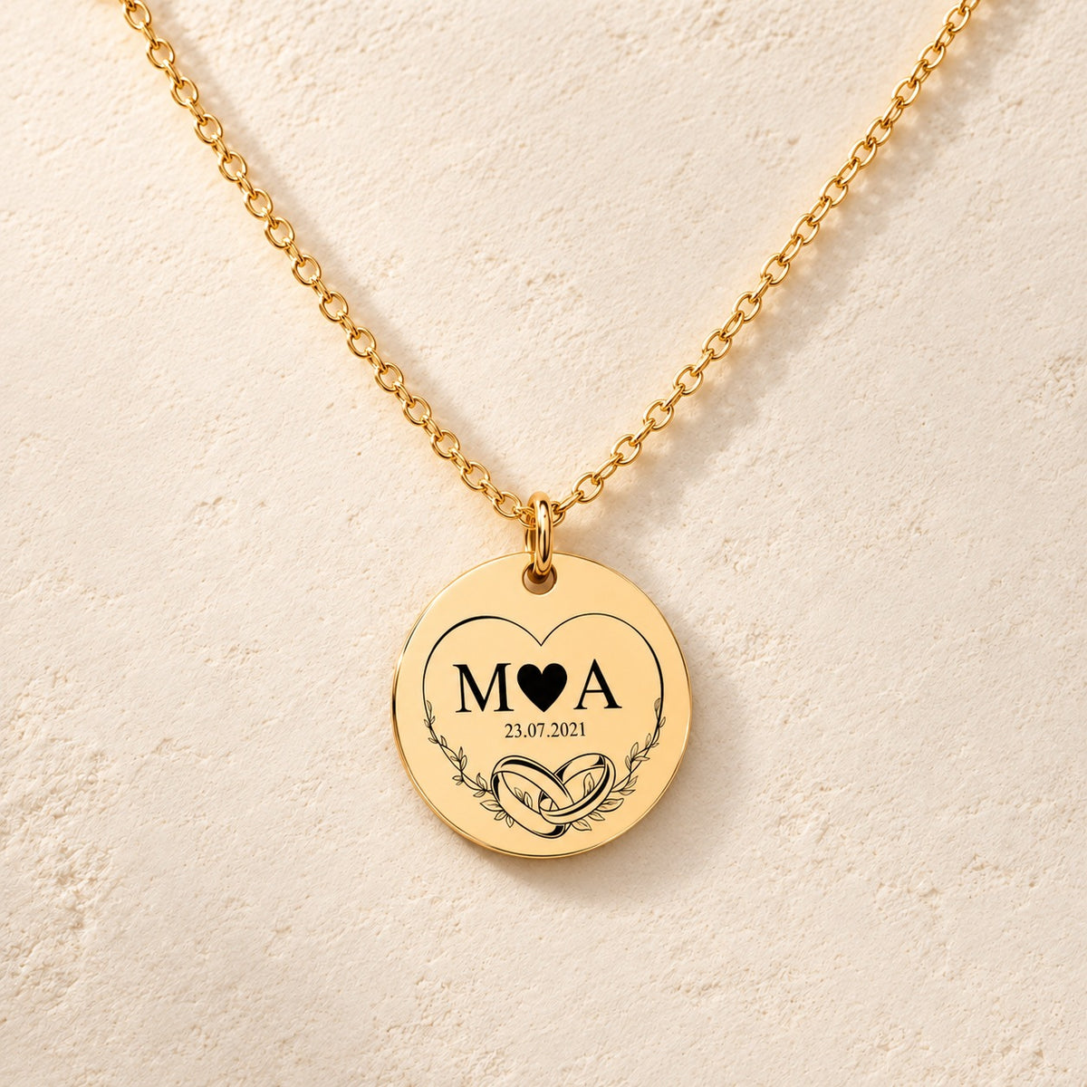 Collier initiales entrelacées Eternal Love personnalisé doré, médaille ronde gravée avec initiales M et A, deux alliances et date 23.07.2021, sur fond ivoire.