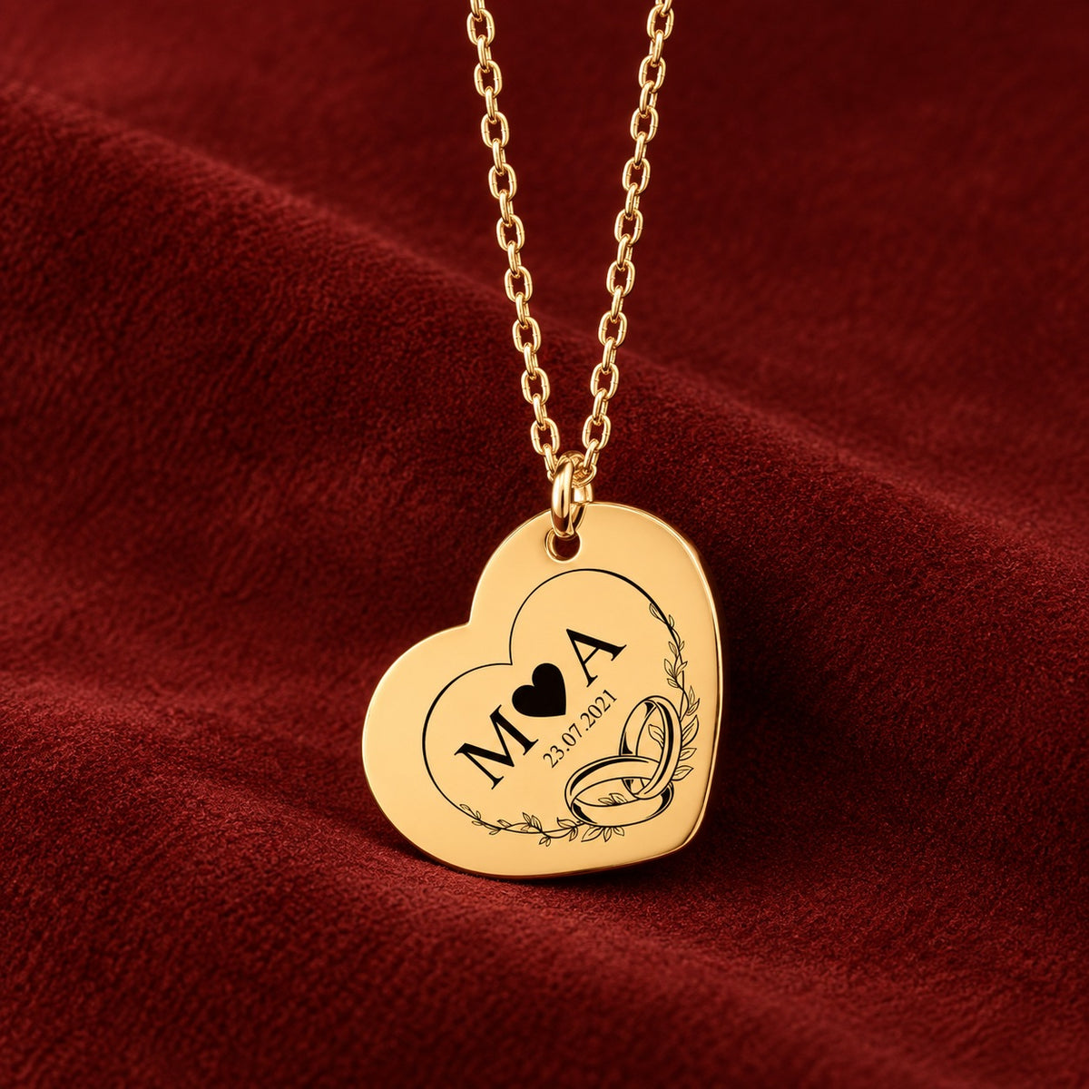 Collier Timeless Bond cœur doré avec initiales M et A, date et alliances entrelacées, sur suédine rouge sanguine.