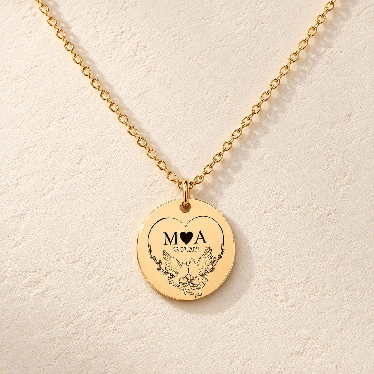 Collier Initiales Entrelacées personnalisé doré, médaille ronde gravée avec initiales M et A, deux colombes et date 23.07.2021, sur fond ivoire.