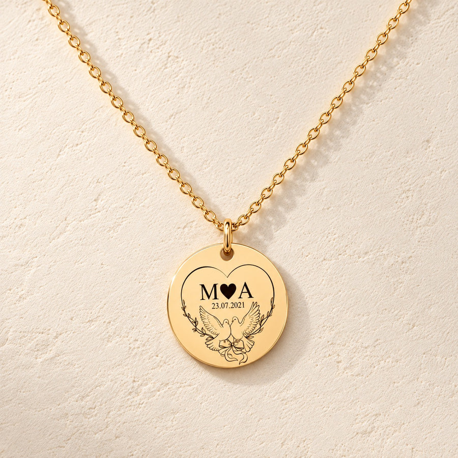 Collier Initiales Entrelacées personnalisé doré, médaille ronde gravée avec initiales M et A, deux colombes et date 23.07.2021, sur fond ivoire.