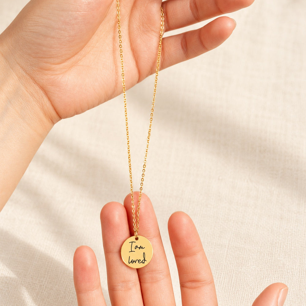 Collier Médaille Affirmations doré tenu entre les doigts, détail de la médaille et de sa gravure.