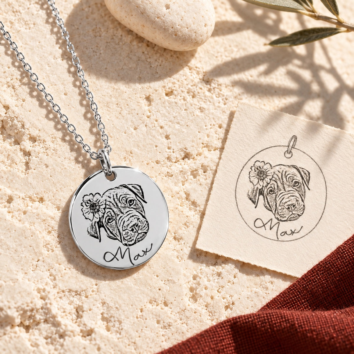 Collier portrait de chien Max argenté sur pierre claire au soleil, avec dessin du pendentif, ombres d’olivier et étoffe rouge sanguine.