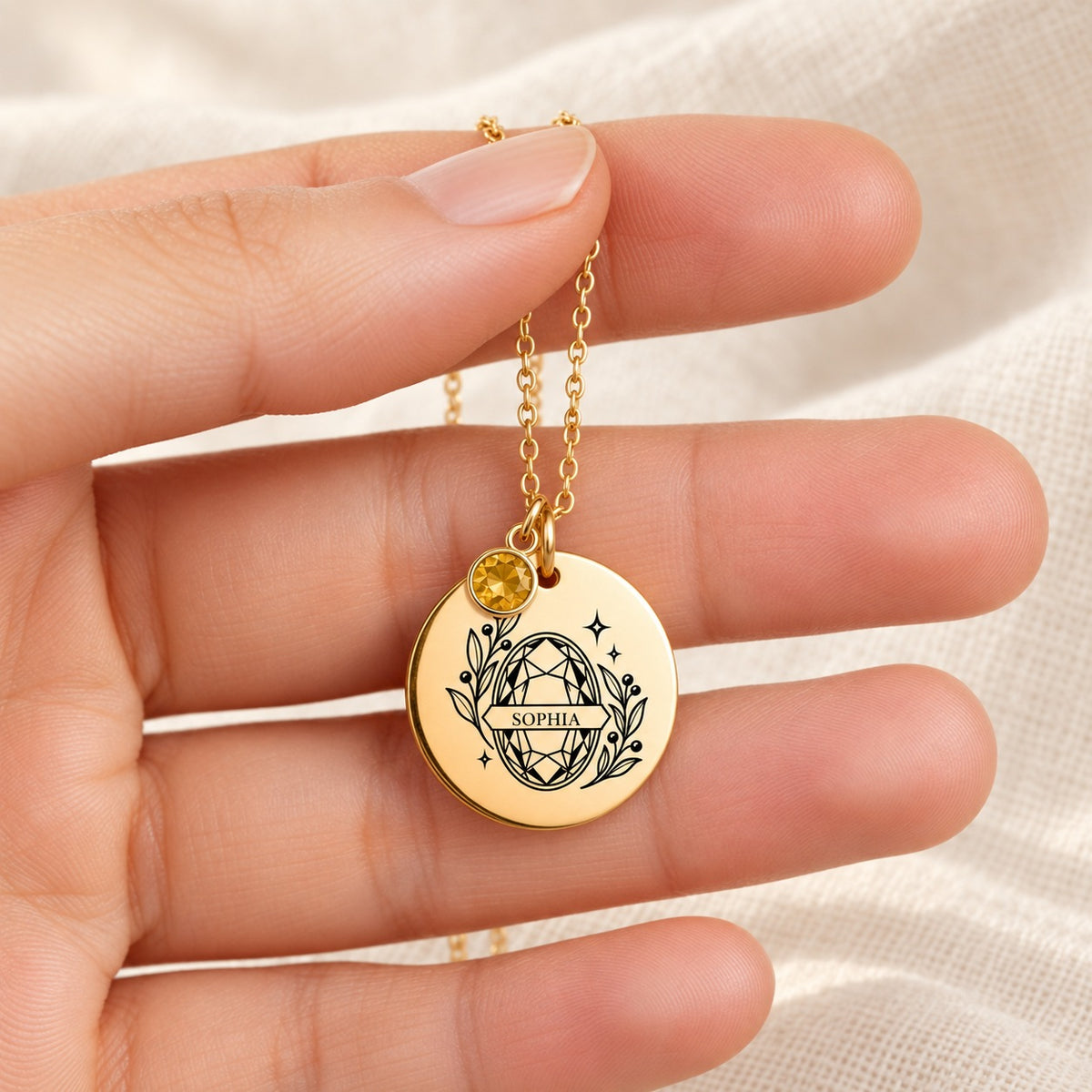 Collier Prénom et Pierre de Naissance doré tenu entre les doigts, détail de la médaille et de sa gravure.