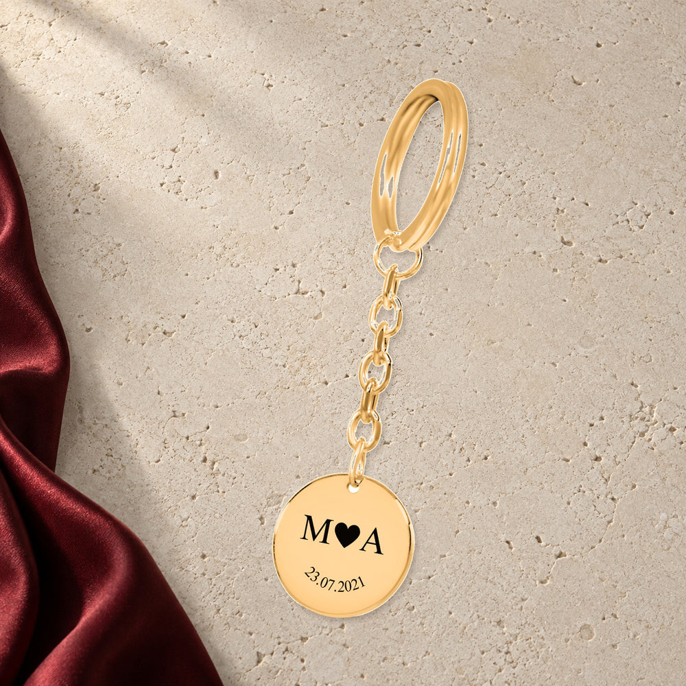 Porte-clés Initiales Entrelacées Personnalisé (Etched In Time) en finition dorée sur pierre claire, avec étoffe rouge sanguine ; exemple de personnalisation.